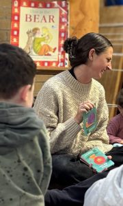 Amber Jackson, creative arts therapist, sits on the floor with preschool children, smiling and holding colourful cards during a group session at Koru Allied Therapies.